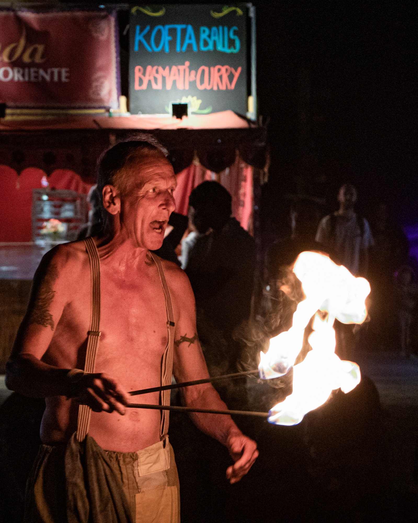 A fire breather exhales a bloom of flame at a night market, neon signs glowing behind him