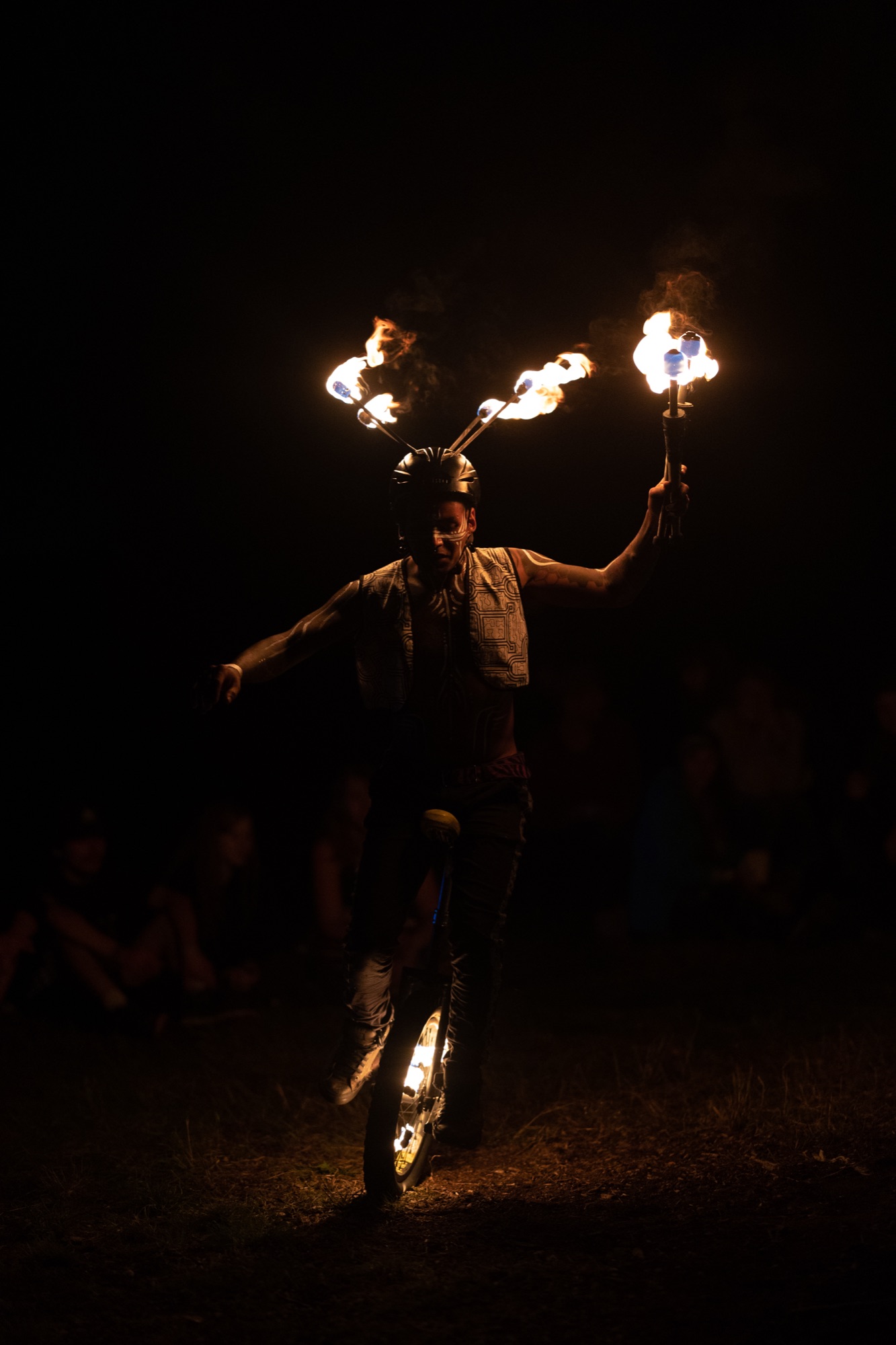A solo fire dancer holds two burning fire fans overhead, body lit only by the flames