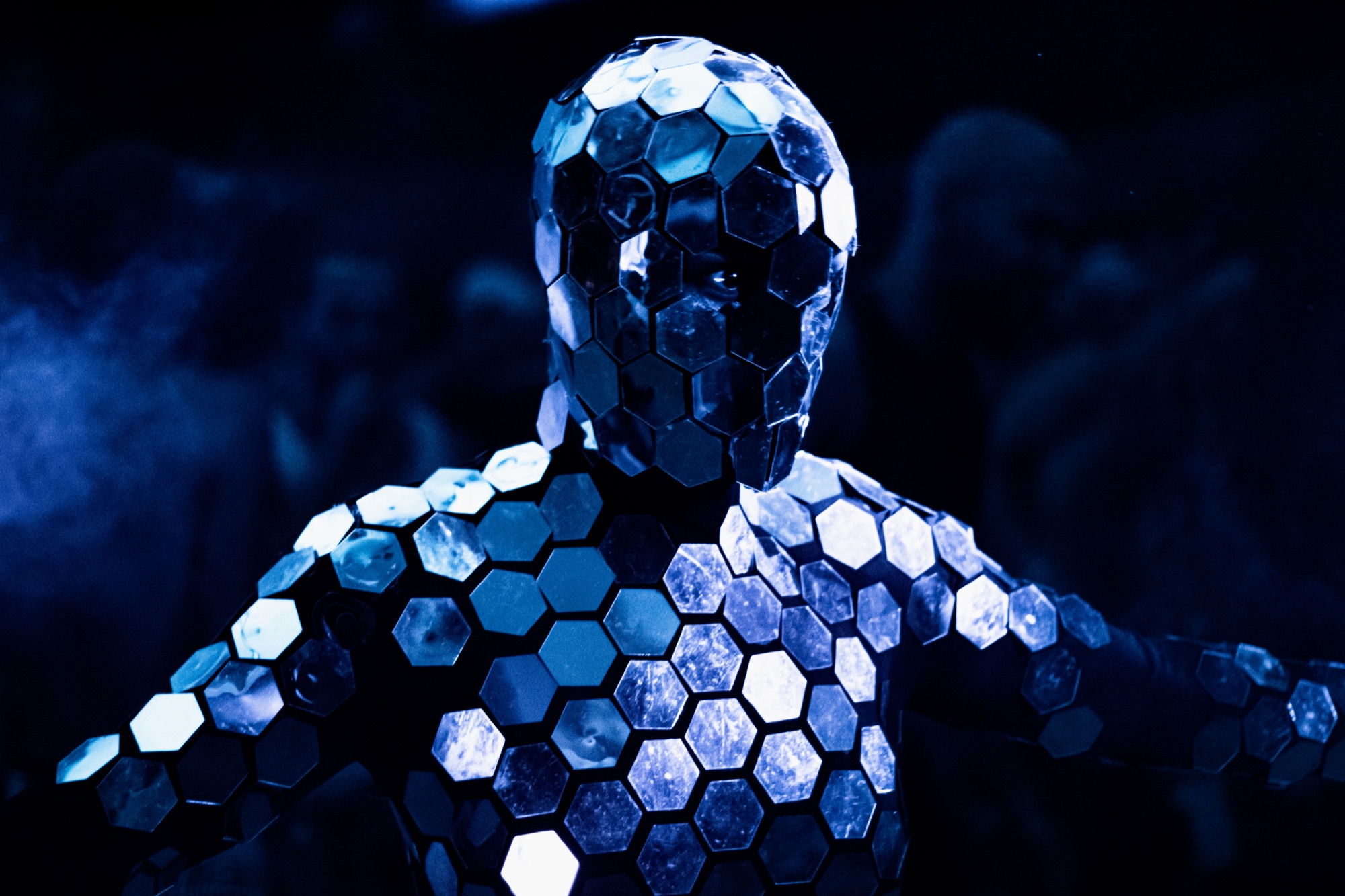 A performer covered in hexagonal mirror tiles, lit blue through drifting smoke