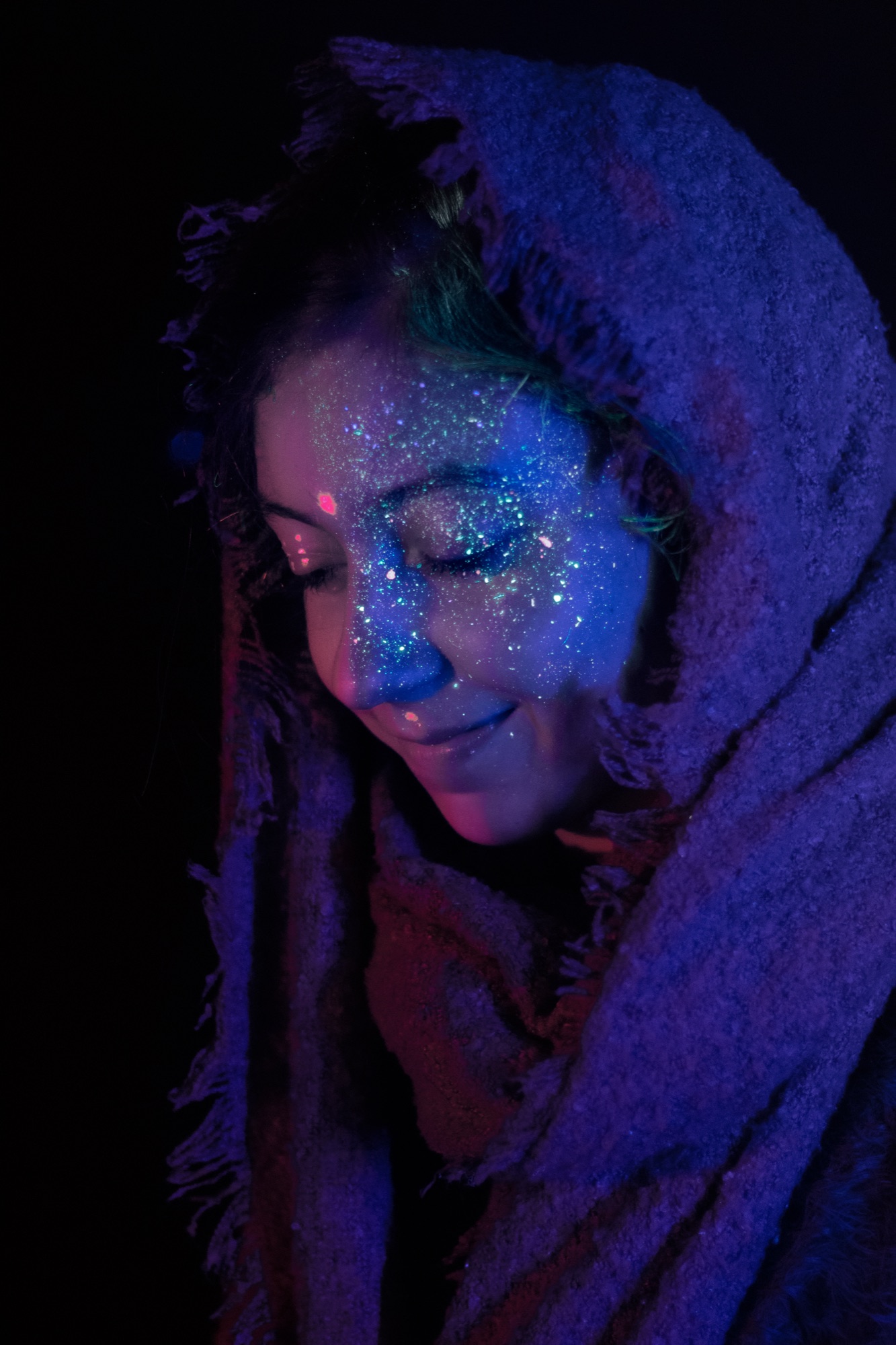 A woman in a hood with glitter face paint, lit in deep purple ultraviolet light