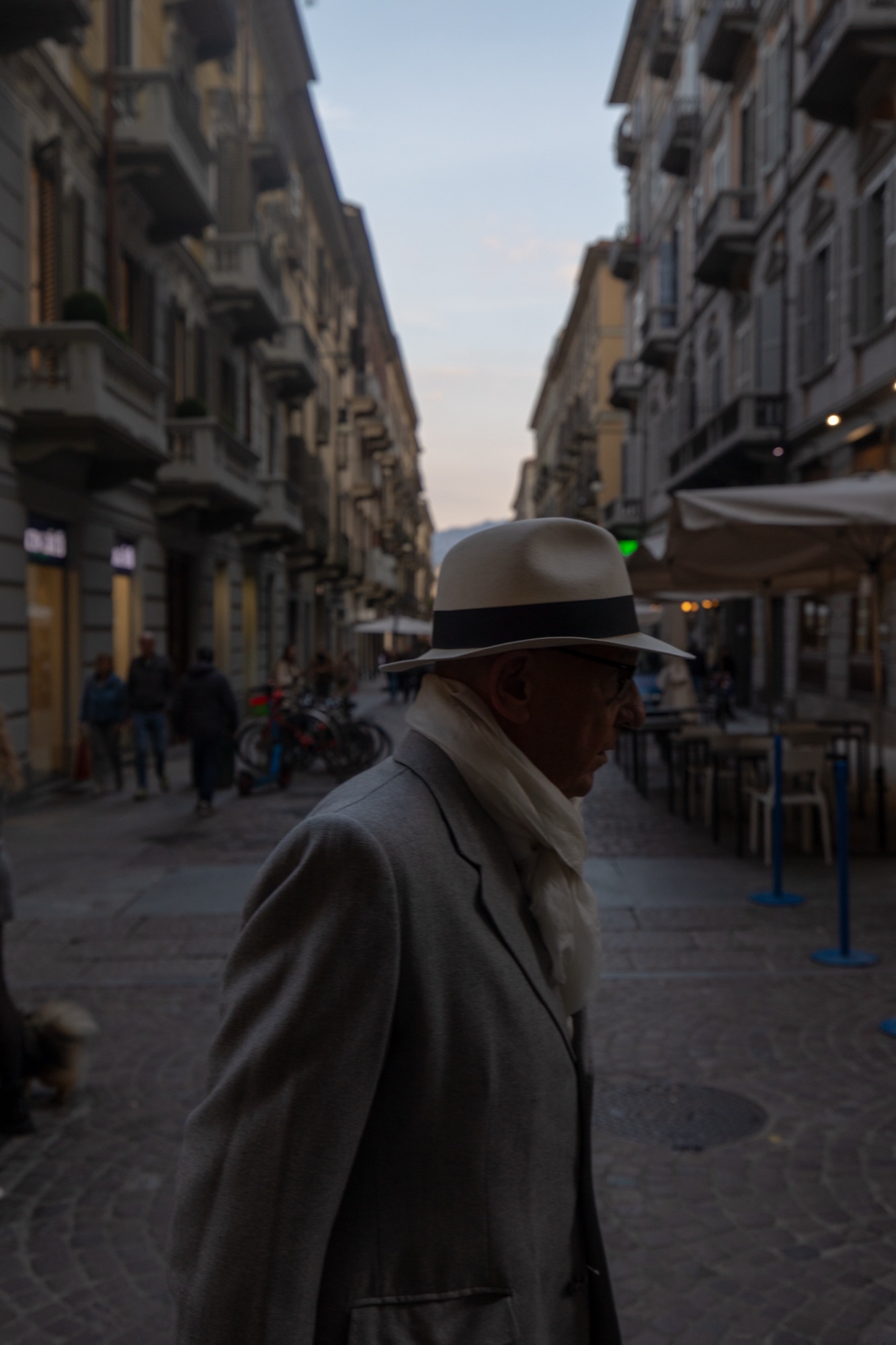 A man in a white hat and suit walking through a narrow European street